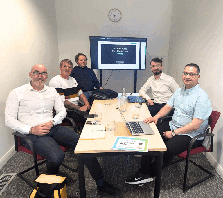 Five people gathered around a meeting table with laptops, notebooks, and water bottles. A screen in the background shows a presentation slide, and a clock is mounted on the wall above it.