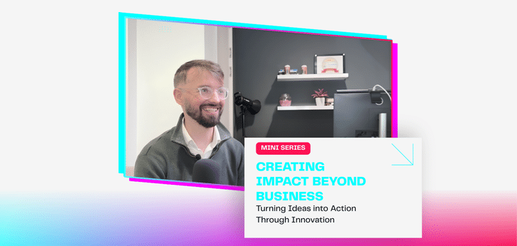 A person sits at a desk in a modern office speaking into a large microphone on a stand. The person wears glasses and a grey zip-up sweater over a light shirt. Behind the desk are a computer monitor, shelves with small decorative items, and a dark grey wall. On-screen text reads: “Artur Grzybowski – Innovation Director, Salford Innovation Forum.